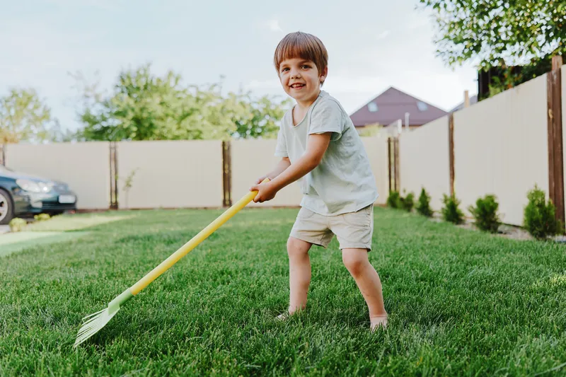 Cheerful boy playing in the garden, digging soil with a yellow rake while smiling under a blue sky, surrounded by green grass and fresh plants.