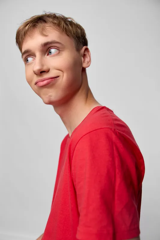 Man in a red shirt with a subtle smirk and sideways gaze, casual studio shot conveying playful confidence and candid charm, clean background, soft lighting, modern lifestyle vibe