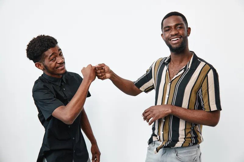 Two young black men with dark skin fist bumping and smiling in casual clothes on white background for friendship, connection, and positive interaction.