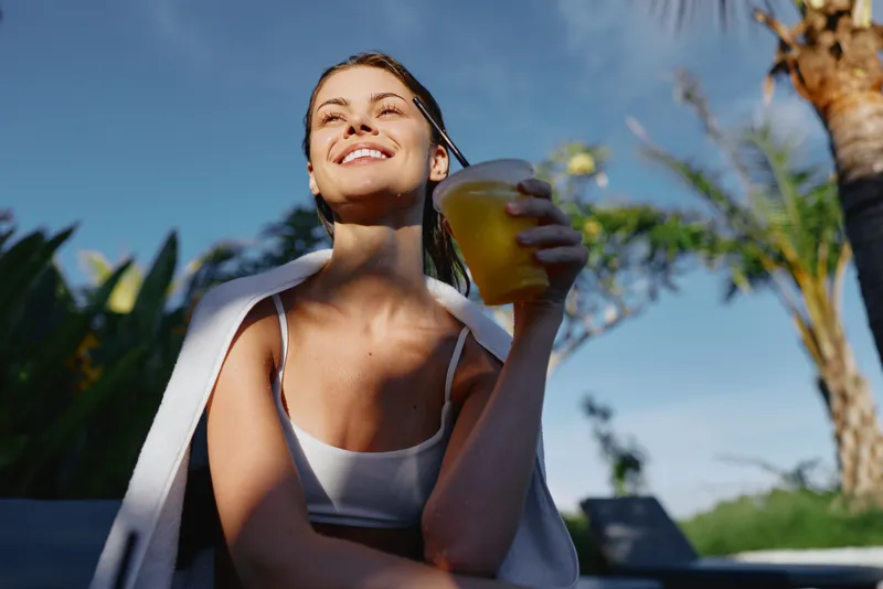 Smiling woman outdoors enjoying a refreshing drink, sunlight on her face, relaxed mood, casual clothing, clear blue sky, tropical plants in background, summer lifestyle