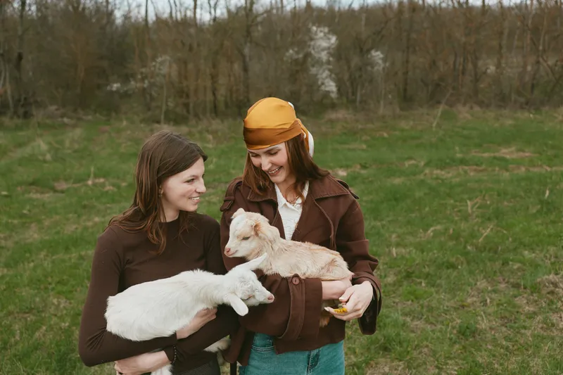 Two young women enjoy a countryside moment holding a white and a brown baby goat in their arms while smiling and standing on a green grassy field during springtime.