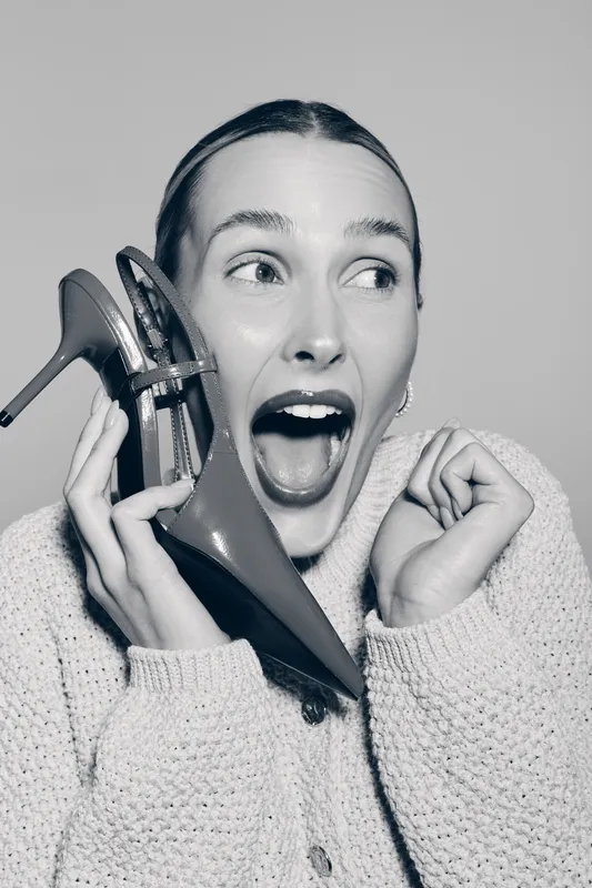 Young white woman with light skin holds a high heel shoe near her face, expressing excitement and surprise. Studio portrait in black and white with casual sweater and slicked hair.