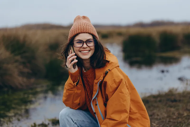 Woman smiling outdoors by a lake, wearing hat and orange jacket, holding a phone to her ear, autumn nature lifestyle with warm colors, soft natural light, blurred landscape background