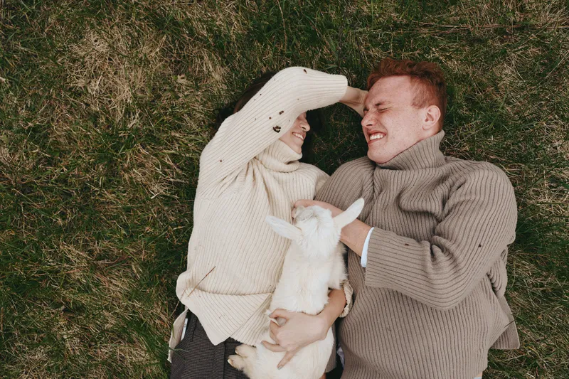 Couple, family, goat, kid, outdoors, grass, happiness, affection, lifestyle, together. A smiling man and woman lie on the grass with a playful goat kid between them, capturing a warm moment of