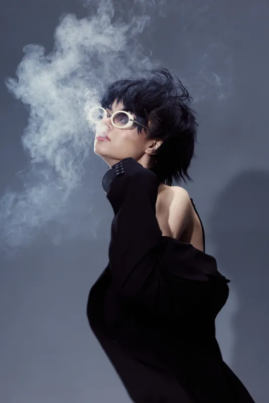 Stylish woman wearing sunglasses exhales smoke against a gray background in a dramatic pose
