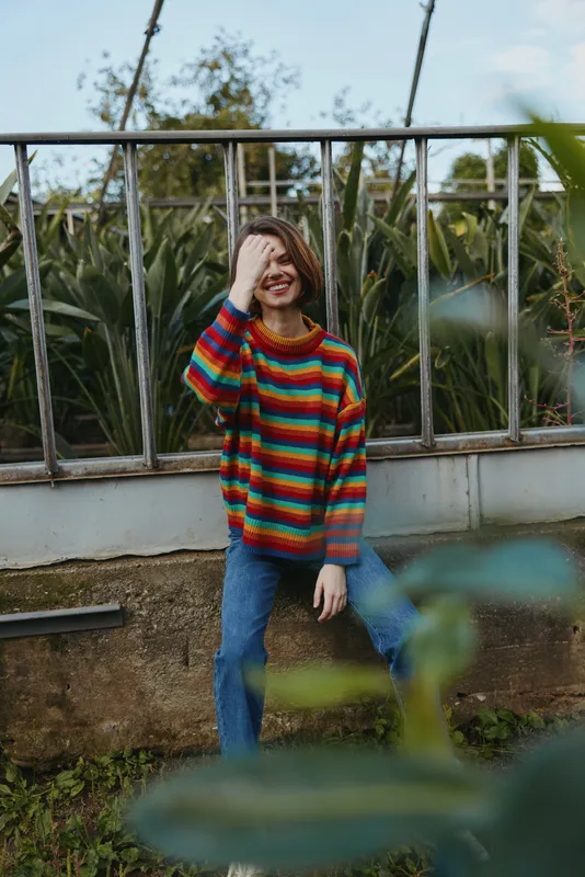 Young woman in a colorful striped sweater sitting in a greenhouse among plants, smiling while covering one eye with her hand, casual outfit and relaxed playful mood for lifestyle.