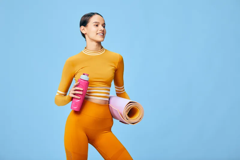 Active woman in bright orange athletic wear holds a pink water bottle and a rolled yoga mat against a blue backdrop, ready for a fitness workout and training session.