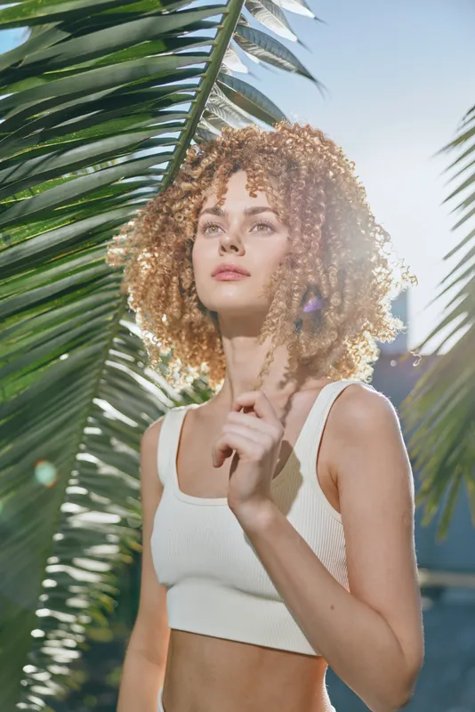 young woman with curly hair wearing a white crop top standing outdoors surrounded by tropical palm leaves under bright sunlight, summer vibe