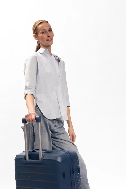 Young woman with a suitcase stands on white background. Casual outfit and relaxed posture create travel and vacation concept. Light and airy studio setting highlights her confident mood.