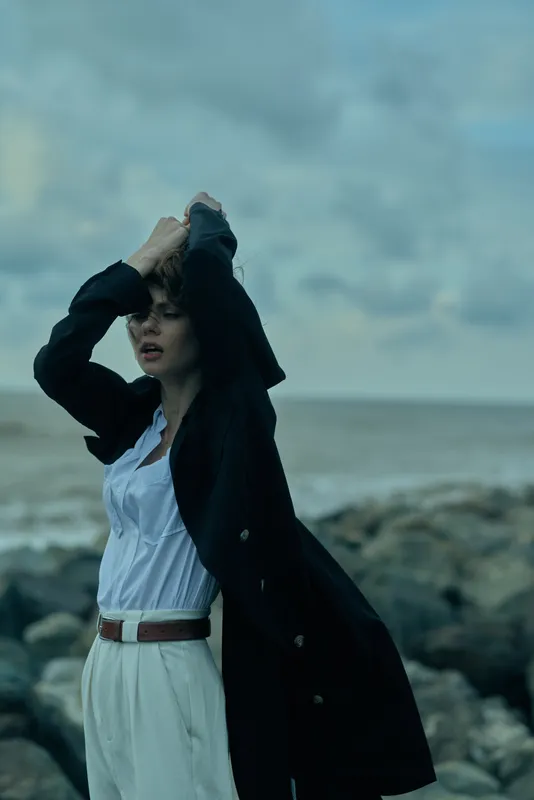 Woman in a white dress and dark coat stands on a rocky coast, sleeves lifted by the wind, creating a cinematic fashion moment against the moody sea and sky.