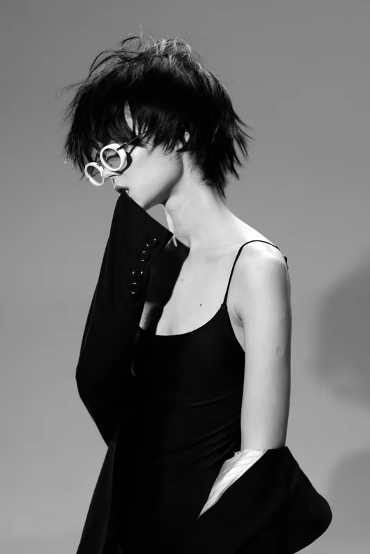 Black and white portrait of a woman with short hair wearing white glasses and a black outfit expressing thoughtful mood.