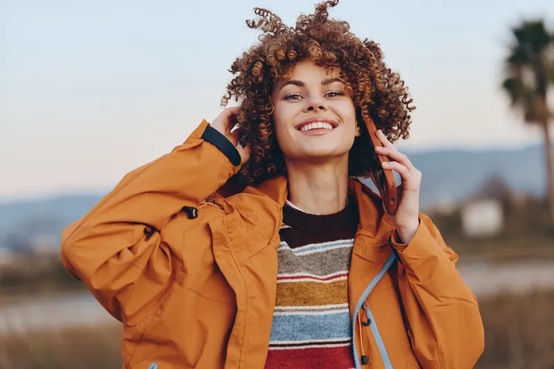 Woman lifestyle outdoors curly hair smile jacket sweater palm landscape, cheerful woman adjusts curly hair in orange jacket, sunlit coast background, relaxed outdoor pose, soft breeze, natural colors