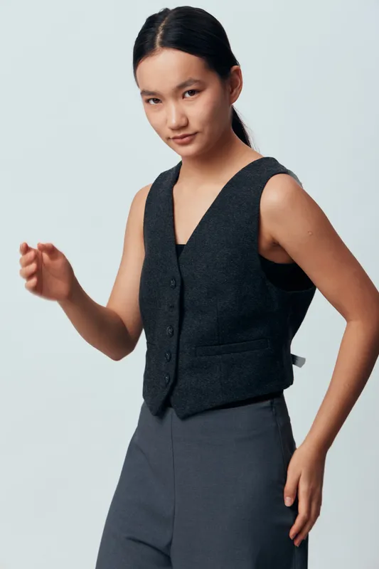 Young woman in dark vest and gray pants posing against light background for fashion portrait, casual style and confidence with natural expression and minimal makeup in studio.
