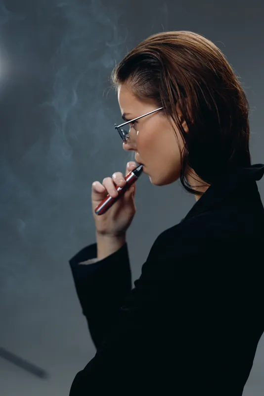 Profile of a young woman vaping indoors with smoke, wearing glasses and dark clothing