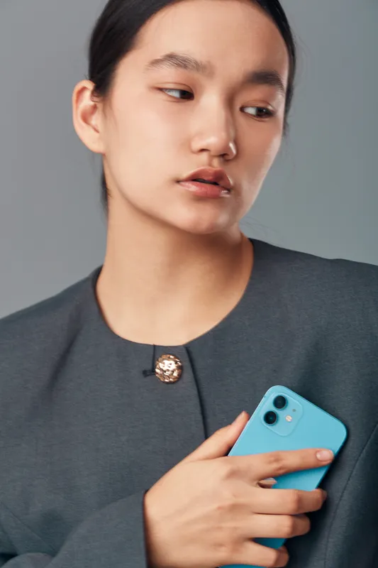 Young woman in gray jacket holding blue smartphone looking away with serious expression on gray background. Portrait, technology, communication, device, casual style, minimalism.