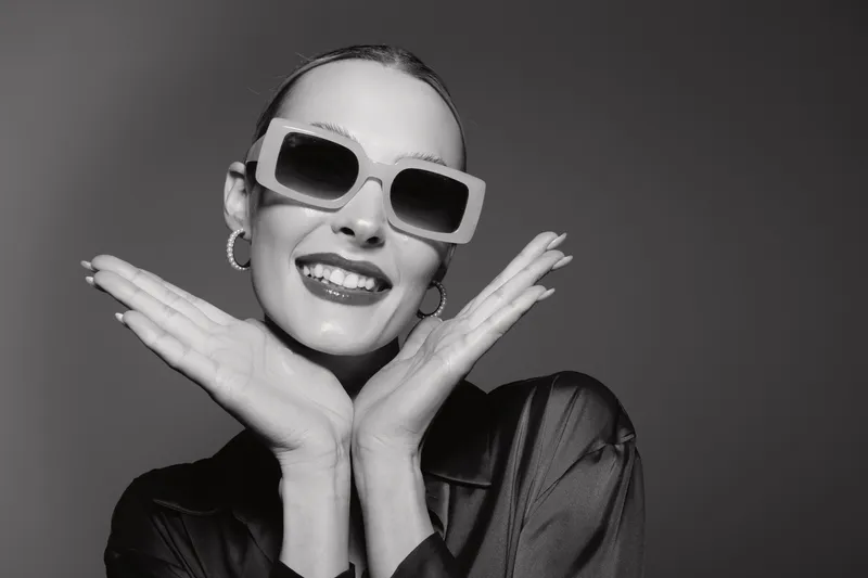 Happy white woman with light skin and stylish sunglasses smiles with hands near face. Studio portrait in black outfit on grey background. Concept of beauty, fashion, and confidence.