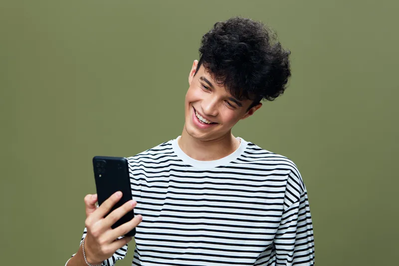 Smiling young man holding smartphone in striped shirt against green background, expressing happiness and casual modern style with curly hair and light accessories.