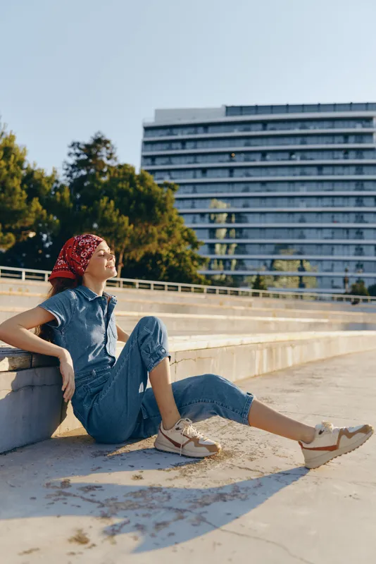 relaxed woman sitting outdoors in sunlight wearing denim jumpsuit and red headscarf near urban park with modern building background