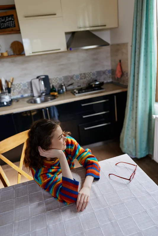Woman with colorful striped sweater sits at a dining table in a cozy kitchen, resting her chin on her hand. Casual home lifestyle scene with soft natural light.