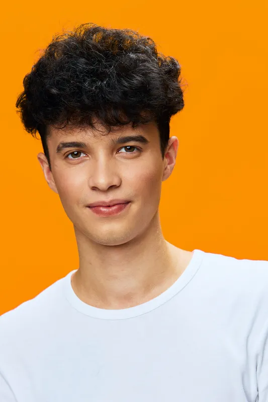 young man with curly hair wearing white t-shirt smiles gently against vibrant orange background, showcasing natural expression and casual style in studio lighting