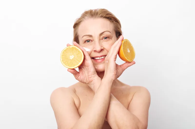 Smiling mature woman holds two halves of fresh grapefruit near face, showing natural skin and healthy lifestyle. Bright studio background highlights freshness and wellness concept for skincare.