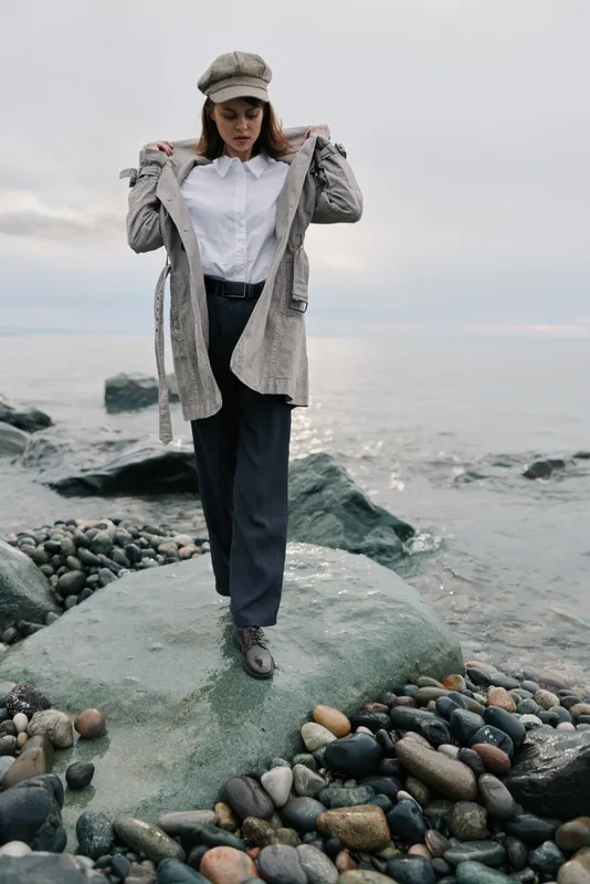 Woman in casual autumn clothes stands on rocky shore near calm sea under cloudy sky. She wears hat, white shirt, cardigan, and dark pants. Concept of nature, fashion, and peaceful mood.