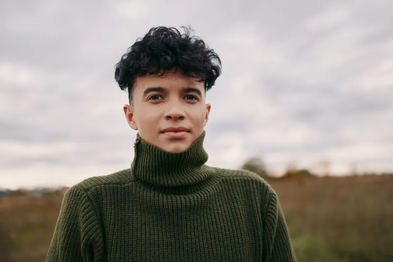 Portrait of a young person with dark curly hair wearing a chunky olive green turtleneck in an open field under soft overcast light, expressing authenticity and credibility.