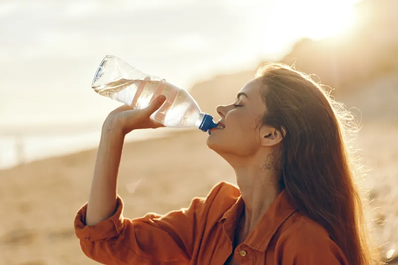 Young woman with long brown hair drinks water from a plastic bottle outdoors during golden hour, enjoying a refreshing moment in warm natural light.
