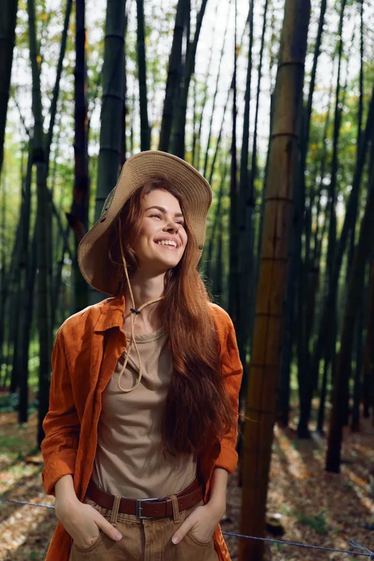 Woman smiling in straw hat in a bamboo forest, redhead traveler wearing casual jacket and knit top, hands in pockets, enjoying natural light and peaceful outdoor portrait.