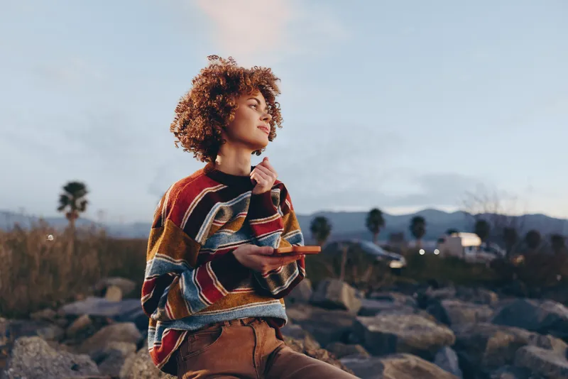 Woman outdoor lifestyle, curly hair, striped sweater, warm sunset light, standing on rocks, relaxed casual pose, soft breeze, scenic landscape, natural colors, calm mood, thoughtful smile