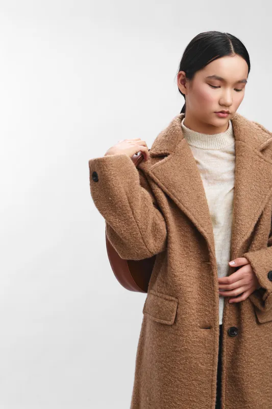 asian woman wearing a warm camel coat stands against a neutral backdrop, adjusting her lapel with quiet confidence, exuding contemporary style, grace, and subtle sophistication in a fashion oriented