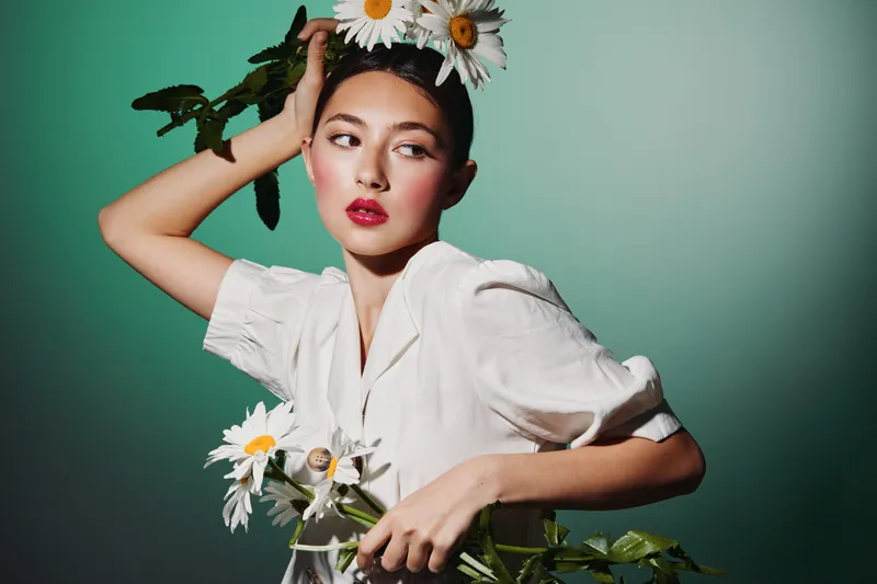 Fashion model wearing white blouse posing with daisy flowers on her head and in hand against gradient green background. Concept of creative beauty and floral fashion styling.