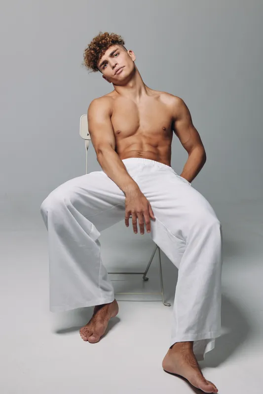 Young man with muscular body shirtless sitting on chair in relaxed pose with white loose pants against plain gray background for fitness health and lifestyle concept.