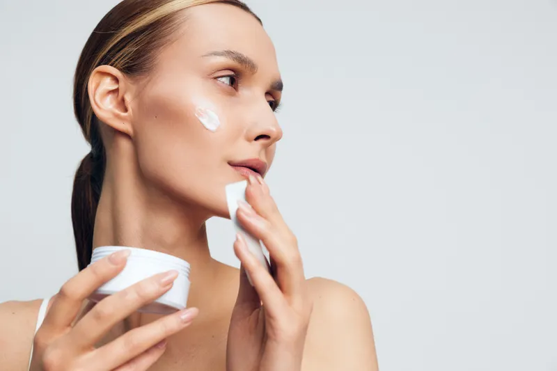 Close-up of a woman applying white face cream on her cheek, on a soft gray background, highlighting skincare routine, moisturization, wellness, and self-care with calm lighting.