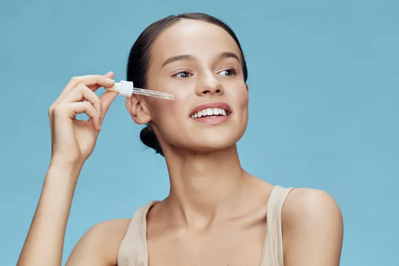 Young woman applying skincare serum with dropper on her face, smiling, against a soft blue background, emphasizing healthy skincare routine and beauty