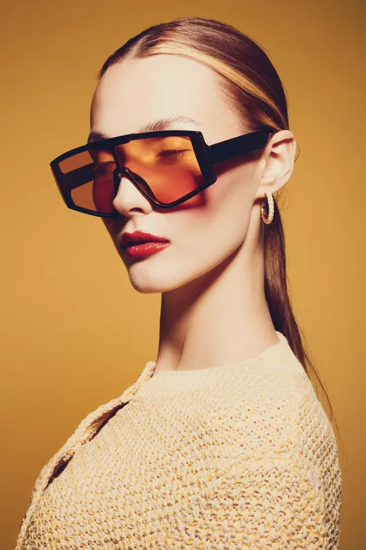 Stylish woman with sleek hair wears oversized orange sunglasses and gold hoop earrings. She poses confidently against a warm yellow background. Concept of fashion, elegance, and modern style.