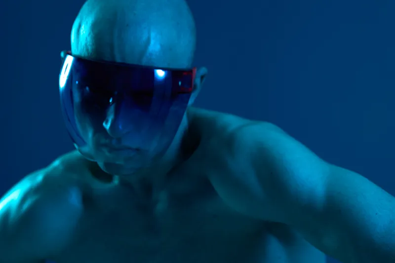 A focused man in a blue light wearing futuristic glasses, showcasing a determined expression and strong physique. Captivating atmosphere highlighting strength and technology.