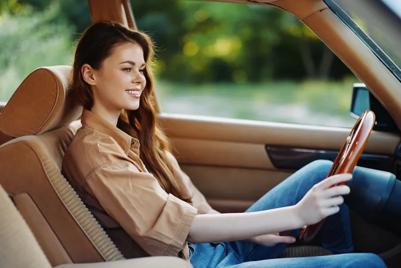 Young woman smiling while driving a classic car in a sunny outdoor setting, embodying joy and freedom in a stylish brown outfit.