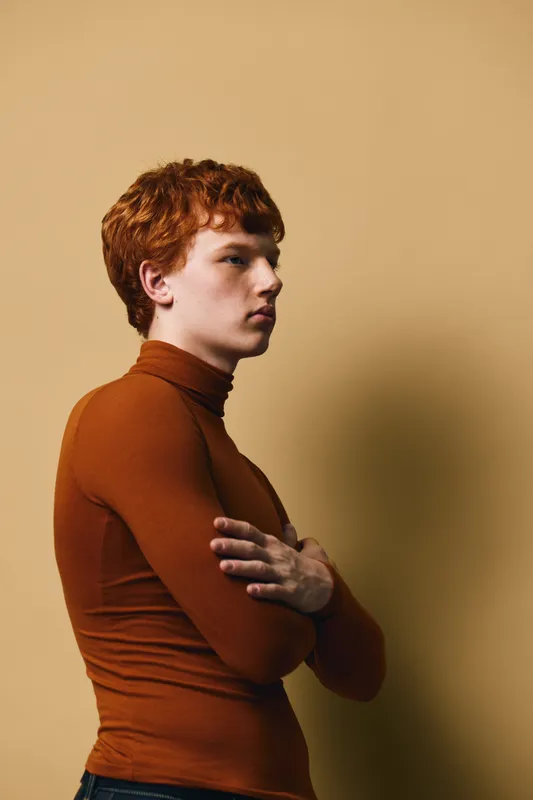 Young man with red hair wearing brown turtleneck standing sideways with folded arms against beige background in thoughtful pose for fashion portrait or casual style.