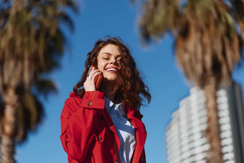 Young woman with curly hair smiles happily while talking on a smartphone outdoors on a sunny day wearing a red jacket and white shirt with palm trees in the background.