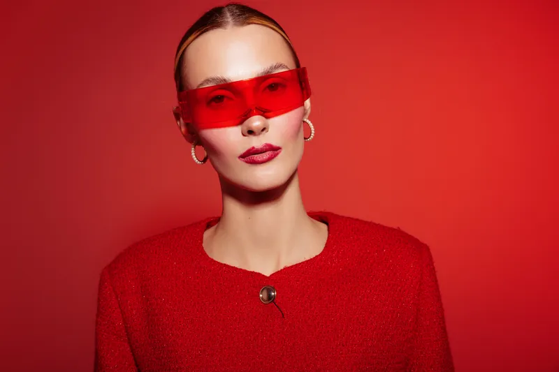 Stylish woman in red jacket and red translucent sunglasses poses against red background. Fashion portrait with bold makeup and sleek hairstyle. Concept of modern style and confidence.