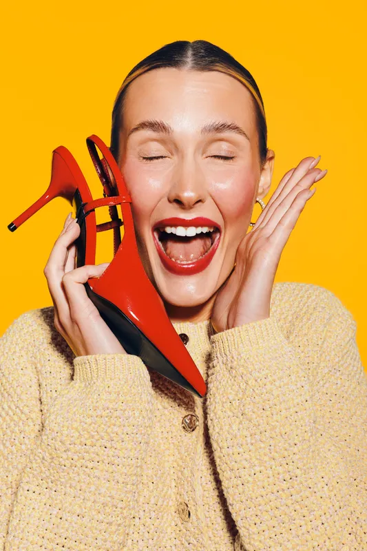 Happy woman with red high heels holds shoes near face, eyes closed and mouth open in excitement. Bright yellow background and cozy beige sweater create joyful mood.