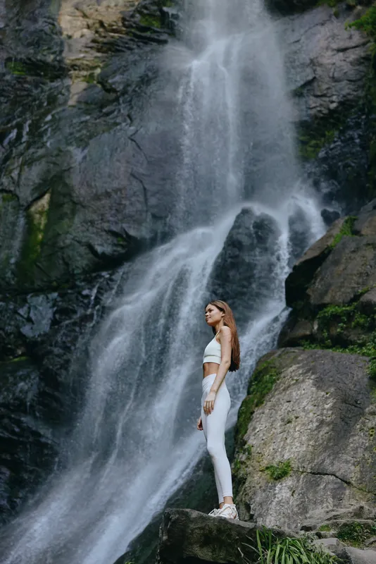woman standing near waterfall in white activewear, natural outdoor setting, rocky landscape, serene atmosphere, fitness and adventure theme