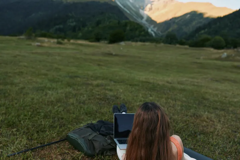 A focused individual sits on grass with a laptop open in a tranquil meadow, mountains in the distance. The scene conveys remote work, creativity, and nature inspired productivity.