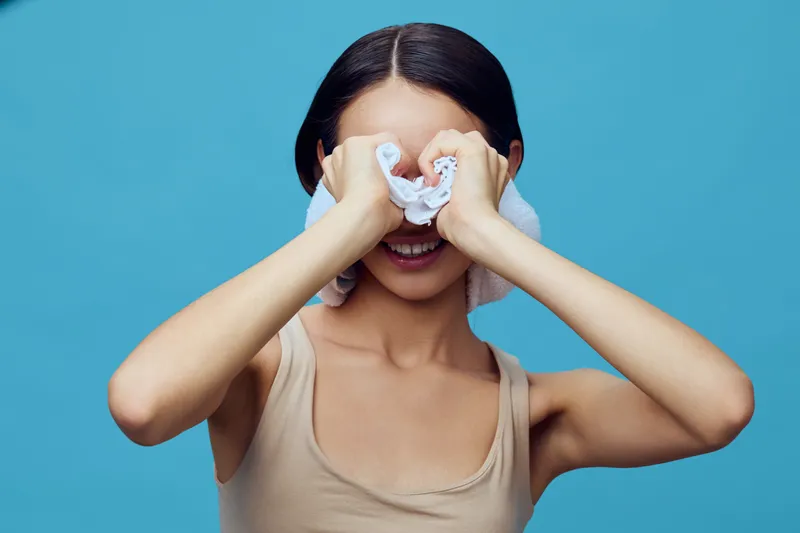 Young woman smiling while playfully covering her eyes with towels against a blue background, showcasing a fun and carefree mood perfect for lifestyle concepts
