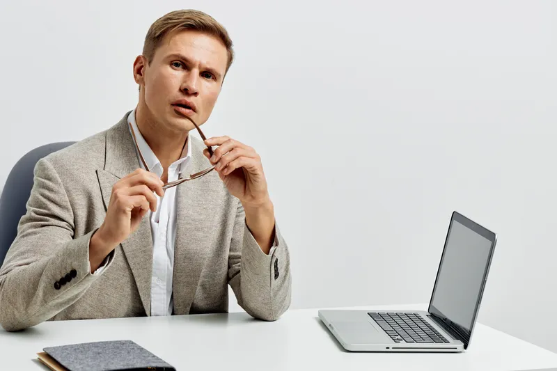 Thoughtful young man holding glasses and looking at camera while sitting at desk with laptop, notepad, and pen in a modern office environment. Focus on concentration, work, and professional atmosphere