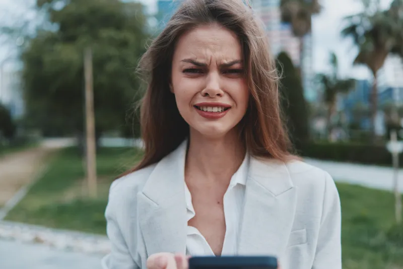 Businesswoman looking distressed while checking smartphone outdoors in an urban park setting, wearing a light blazer and white blouse, conveying frustration and urgency.