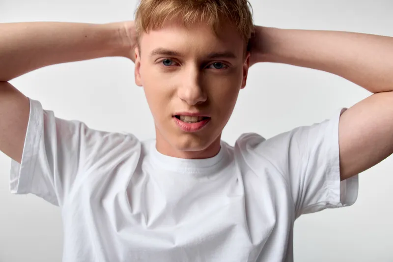 Man in a white t shirt with hands behind the neck and a tense expression, studio shot, casual posture, bold facial focus and contemporary mood
