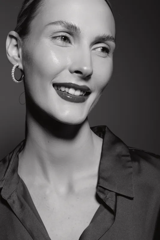 Close-up of a smiling white woman with fair skin and hoop earrings wearing a dark blouse. Studio portrait in black and white with soft lighting highlights natural beauty.
