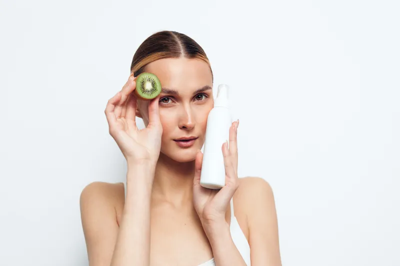 Young woman with clear skin holding a white cosmetic bottle and a slice of kiwi near her face, minimal makeup, clean background, skincare and natural beauty concept